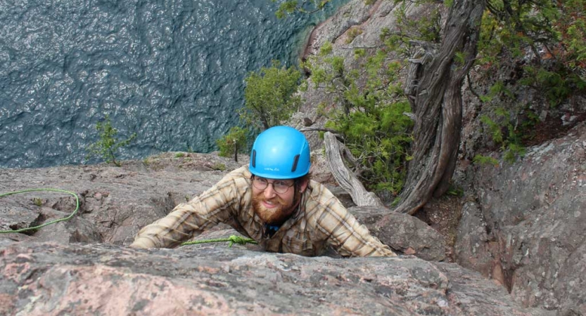 A person wearing safety gear and secured by ropes pauses their climb to look up at the camera. They appear to be high above a body of water. 